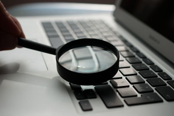 Person using magnifiying glass above keyboard on laptop to symbolize the action of an AI essay checker